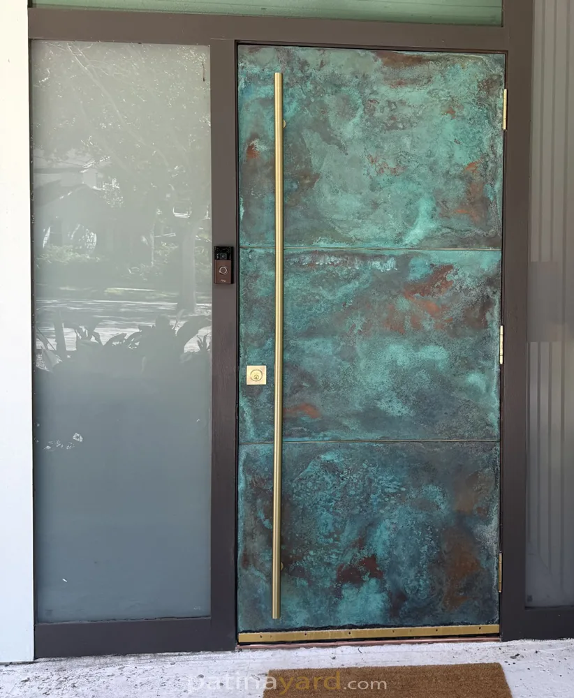 Patina copper front door with a tall brass handle beside a frosted glass sidelight