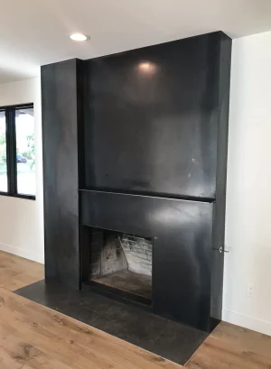 Blackened steel fireplace surround with a shelf ledge and metal hearth in a bright room with wood floors
