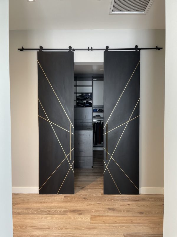 Pair of black stained white oak sliding barn doors with diagonal brass inlays opening to a walk in closet
