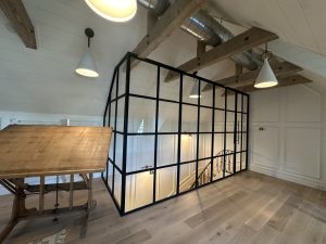 Black metal and glass loft office enclosure with a hinged door under wood beams