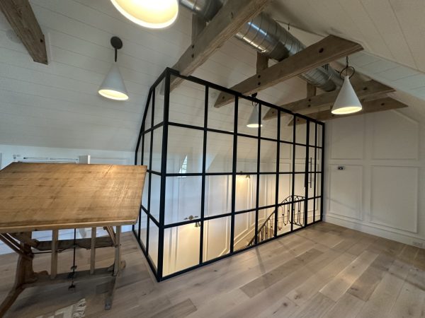 Black metal and glass loft office enclosure with a hinged door under wood beams