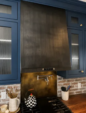 Hot rolled steel range hood with a blackened patina finish over a brass backsplash in a navy kitchen
